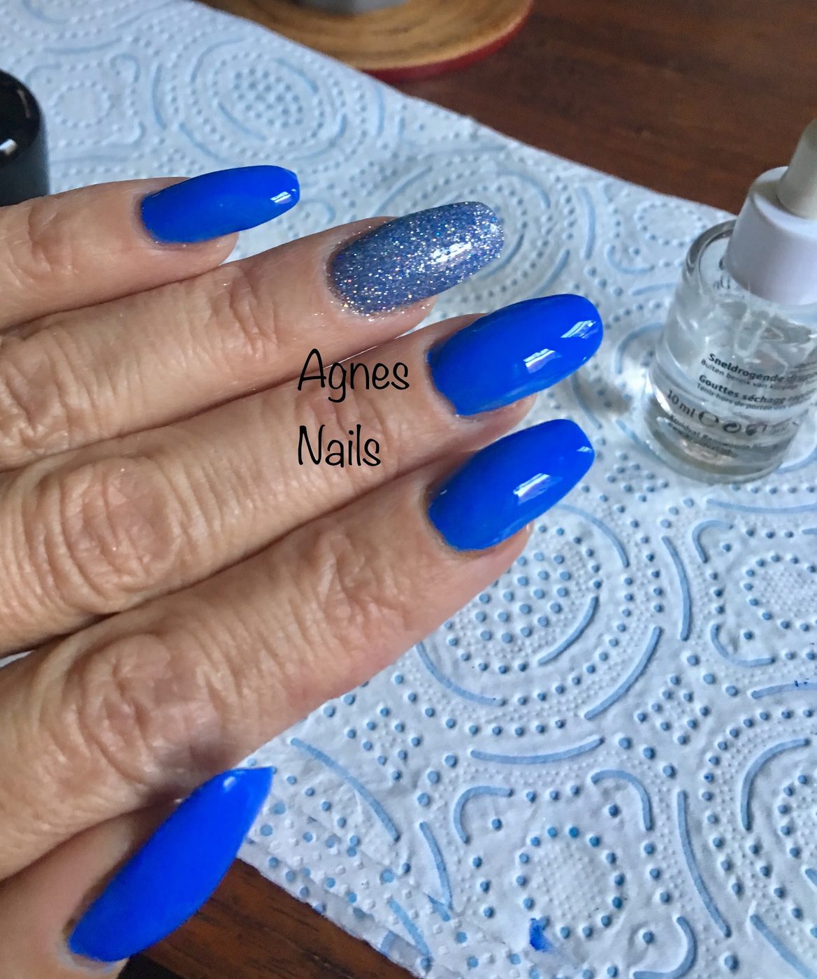 Kobaltblauwe nagels met één glitternagel, hand op een papieren doekje naast een flesje
